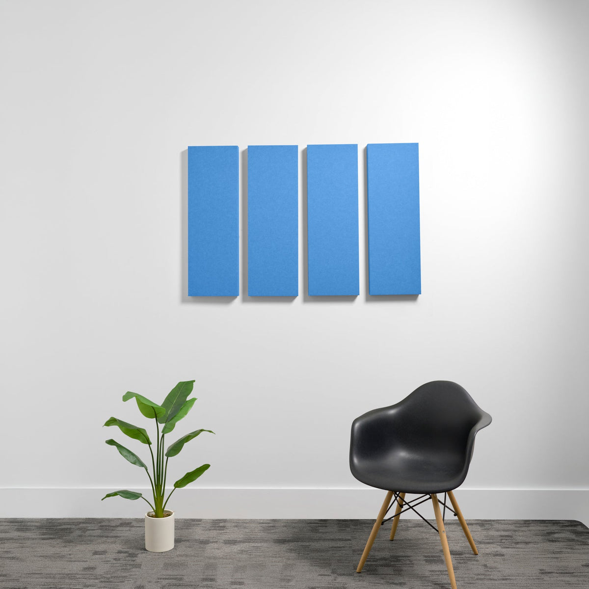 Four blue rectangular acoustic panels mounted on white wall above black chair and potted plant.