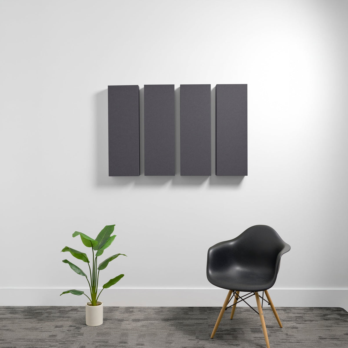 Four dark gray rectangular acoustic panels mounted on wall above modern chair and potted plant.