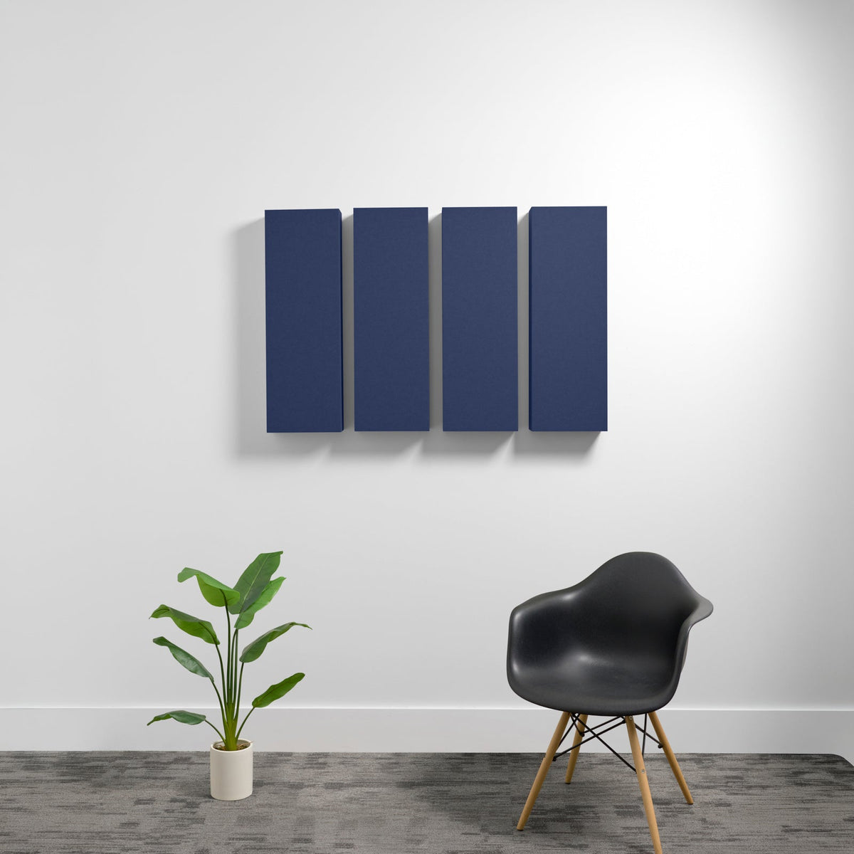 Four dark blue rectangular acoustic panels mounted on white wall above plant and black chair.