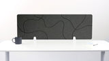 Dark gray desk divider with organic curved line pattern, positioned on white desk with mounting brackets.