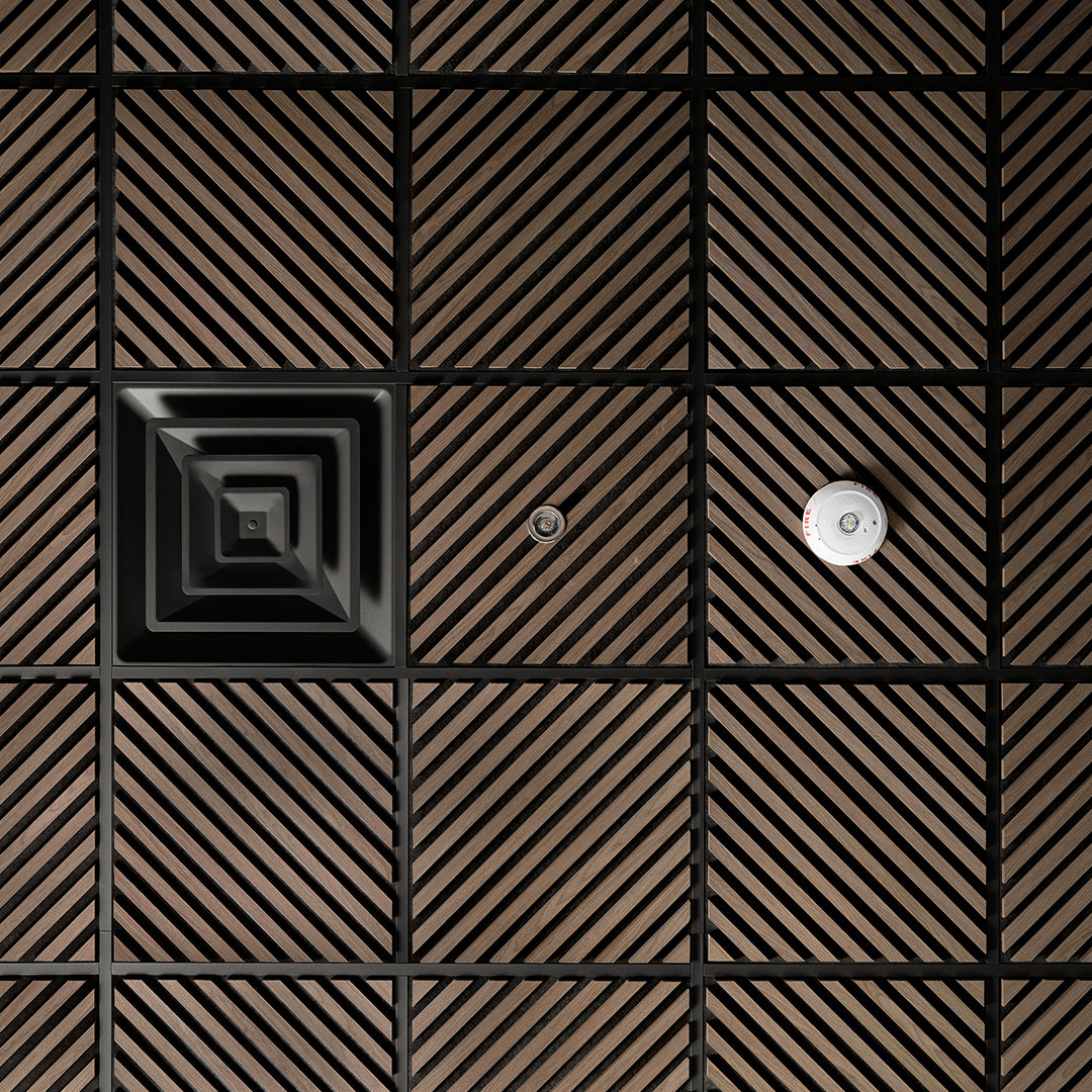 A close-up of a modern wall or ceiling panel featuring a grid pattern of square tiles with diagonal slats in a warm brown tone, separated by black grout lines. One tile has a concentric square design, and another has a small circular metallic fixture.