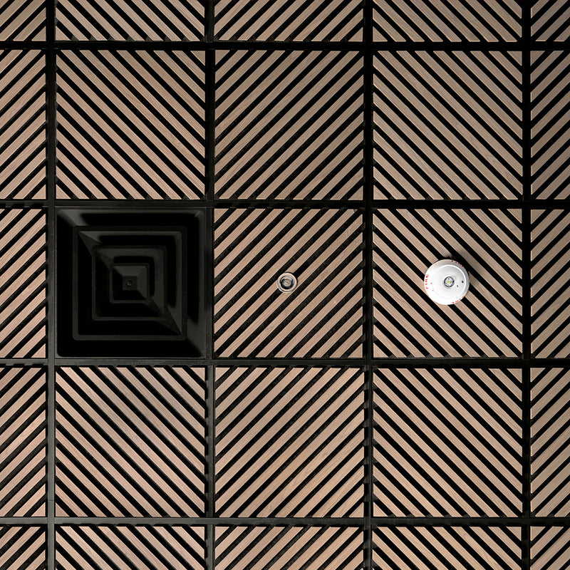 A close-up of a modern wall or ceiling panel featuring a grid pattern of square tiles with diagonal slats in a warm brown tone, separated by black grout lines. One tile has a concentric square design, and another has a small circular metallic fixture.