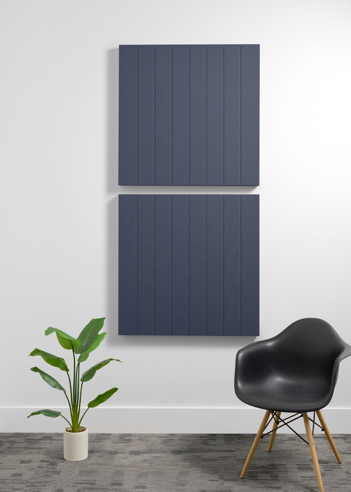 Two dark blue acoustic wall panels with vertical ribbed texture mounted on white wall above potted plant and black chair.