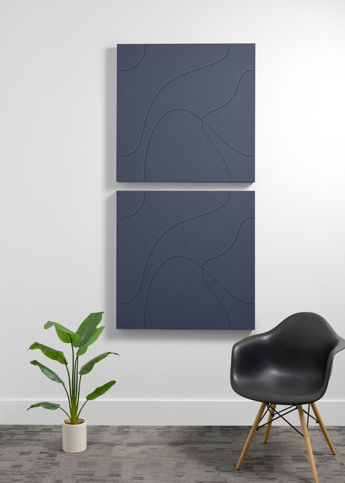 Two stacked dark blue acoustic wall panels with engraved curved line patterns, displayed above a potted plant and black chair
