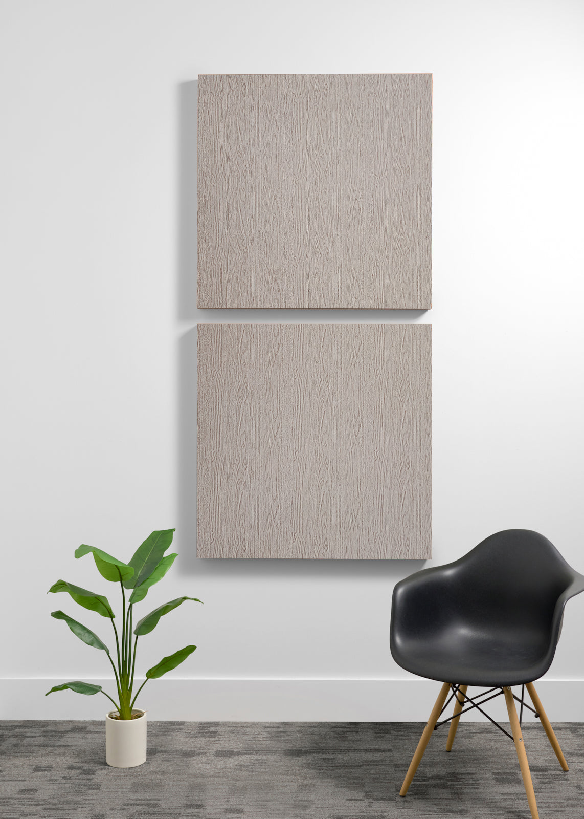 Two square light wood wall art panels hanging vertically on a white wall in a modern room setting, with a black chair and a potted green plant below.