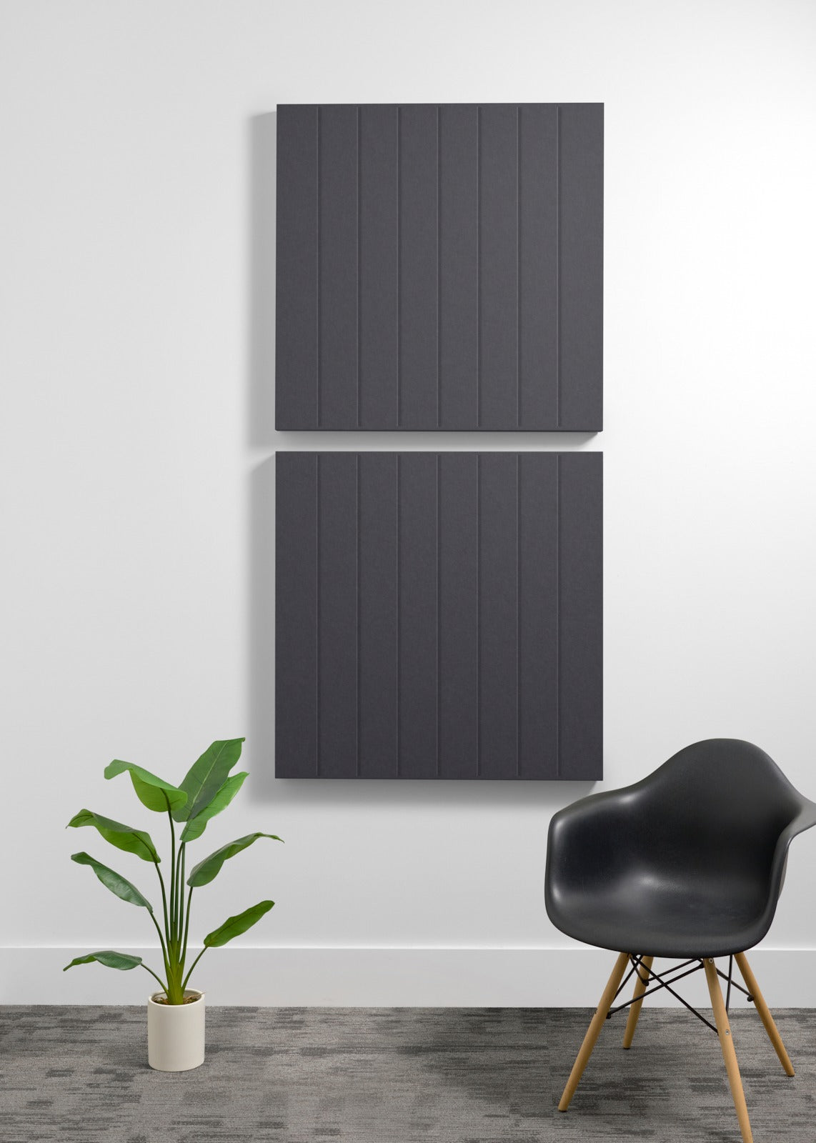 Two dark gray acoustic wall panels with vertical ribbing mounted on white wall above black chair and potted plant.