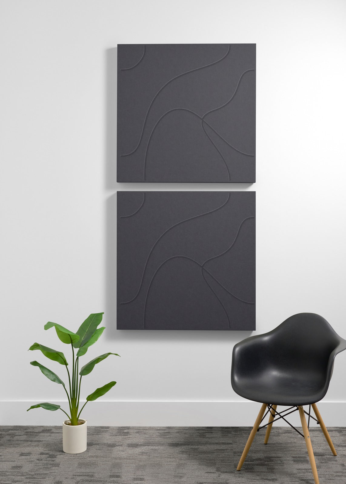 Two stacked dark gray acoustic panels with curved engraved lines mounted on a wall above a chair and plant.