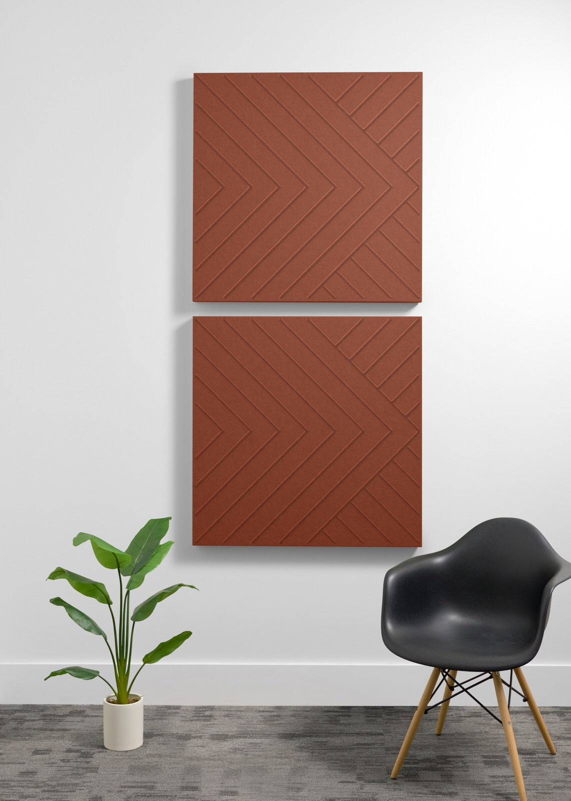 Two terracotta-colored square acoustic panels with chevron engraved pattern mounted on wall above chair and plant.