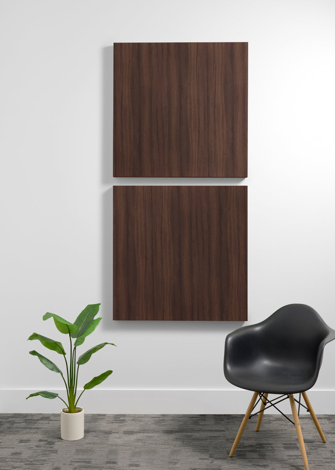 Two square darker wood wall art panels hanging vertically on a white wall in a modern room setting, with a black chair and a potted green plant below.