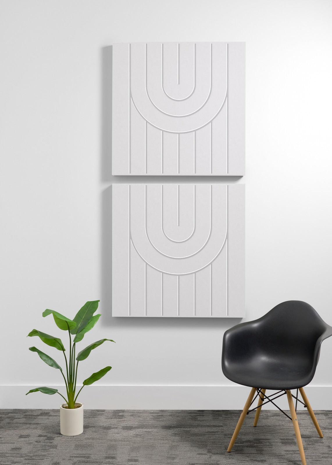 Two white square wall panels with engraved curved line patterns stacked vertically above a black chair and potted plant.