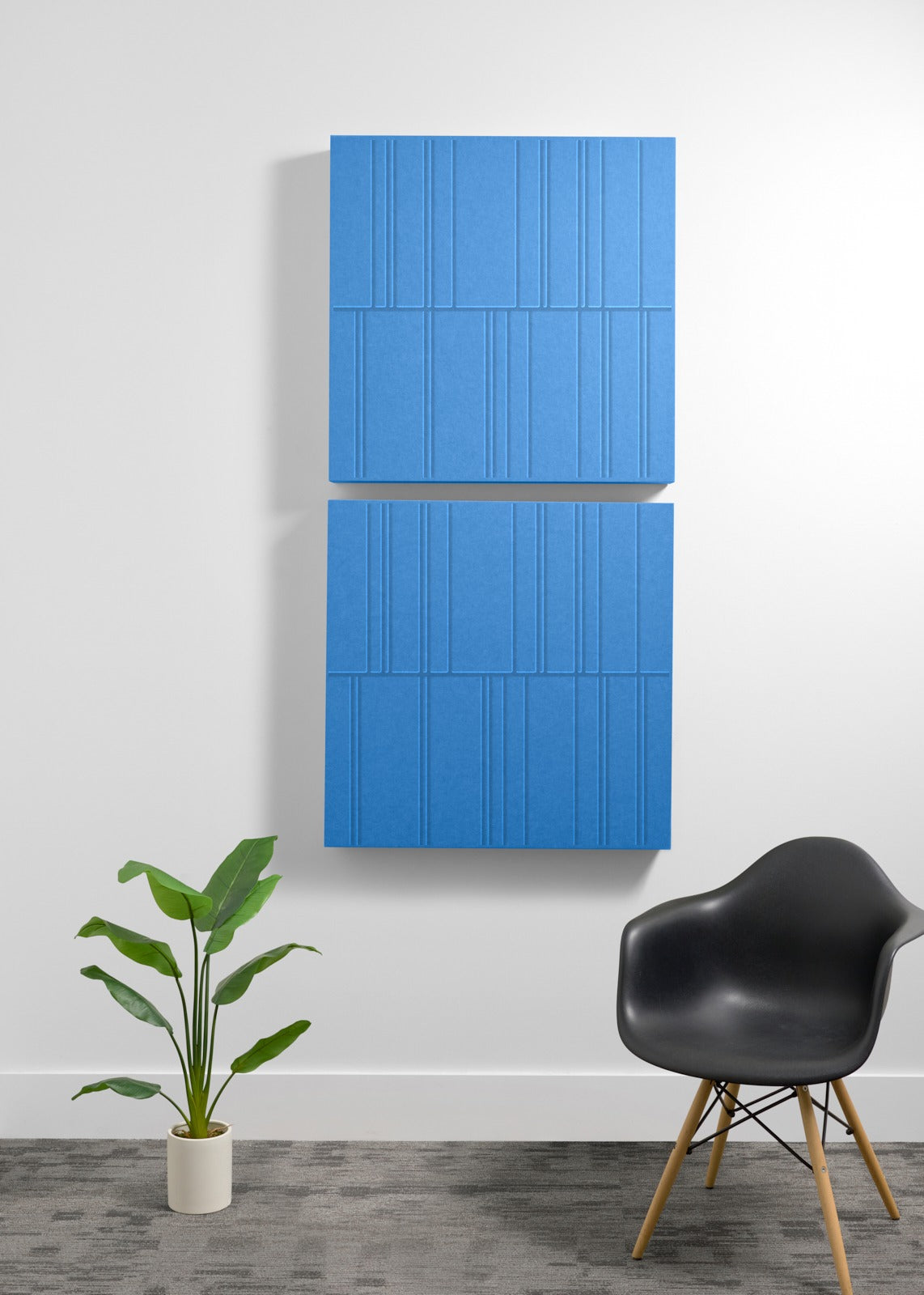 Four blue acoustic panels with vertical engraved lines mounted on white wall above black chair and potted plant.