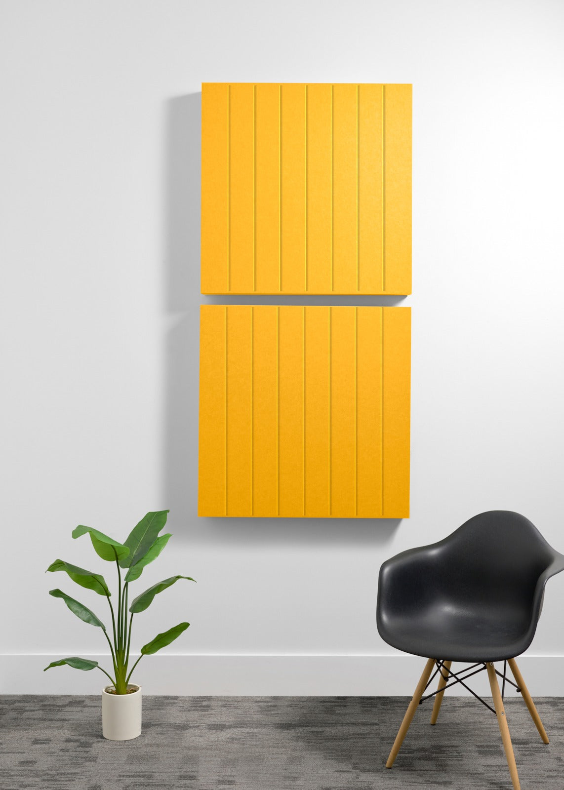 Two stacked yellow acoustic boxes with parallel engraved lines mounted on a wall above a black chair and potted plant.