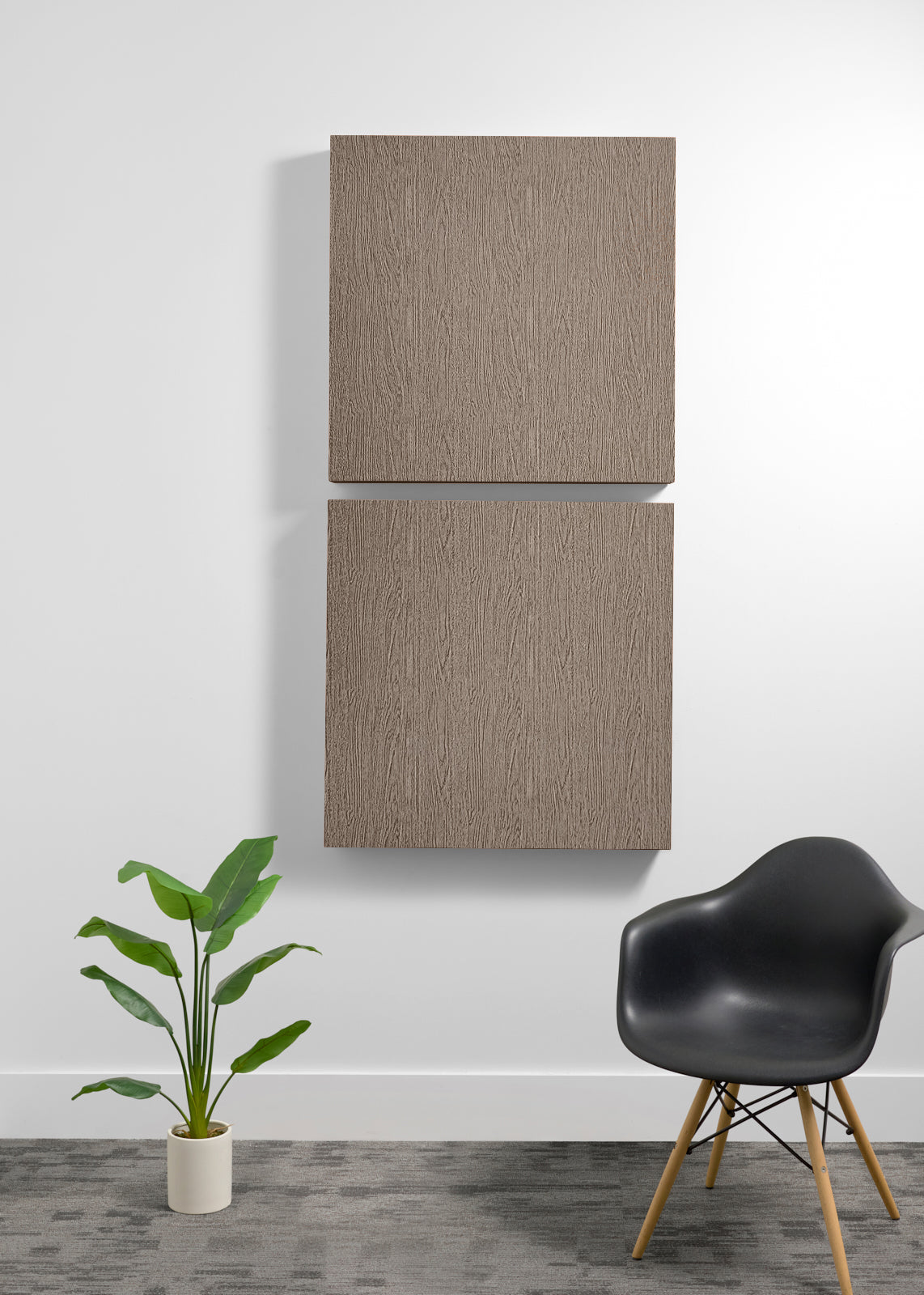 Two square dark wood wall art panels hanging vertically on a white wall in a modern room setting, with a black chair and a potted green plant below.