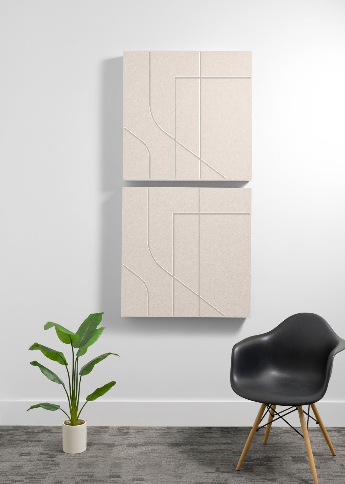 Two ash square wall-mounted panels with engraving on a white wall with a black chair and plant in the foreground.
