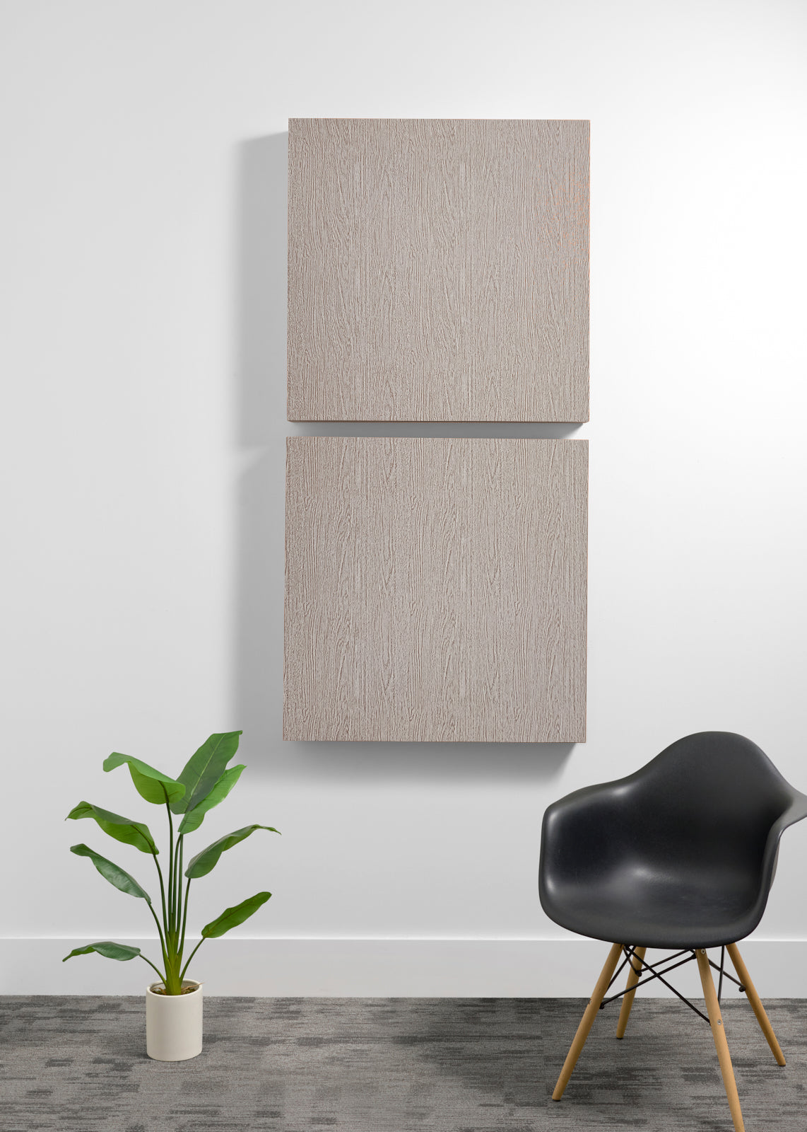 Two square light wood wall art panels hanging vertically on a white wall in a modern room setting, with a black chair and a potted green plant below.