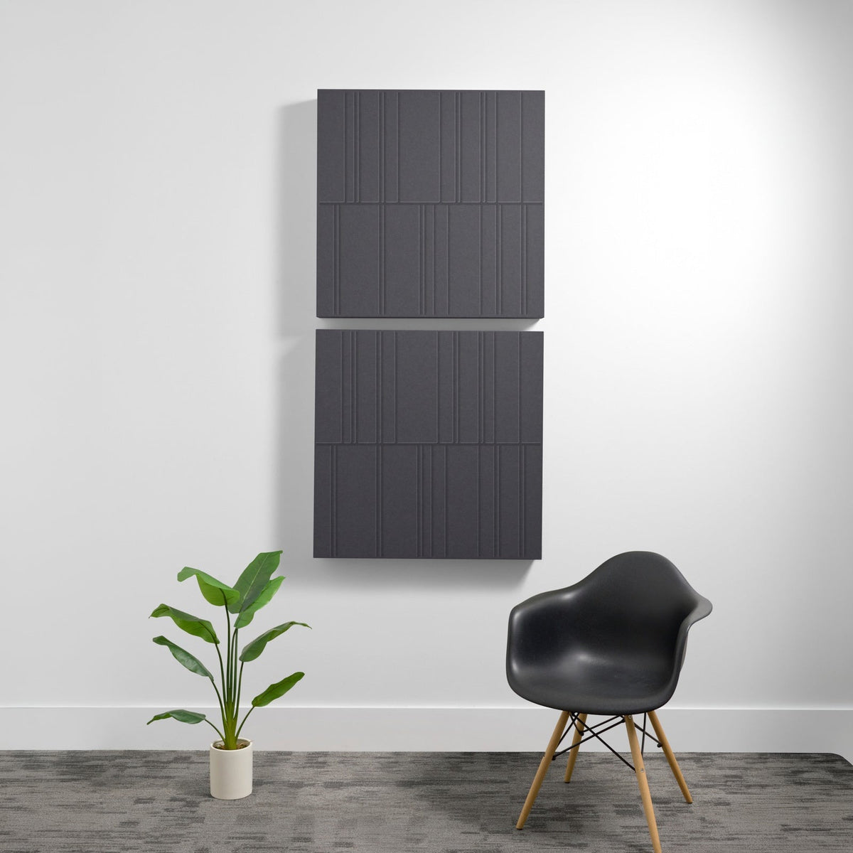 Two stacked dark gray acoustic panels with vertical line engravings mounted on white wall above potted plant and black chair.