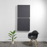 Two stacked dark gray acoustic panels with vertical line engravings mounted on white wall above potted plant and black chair.
