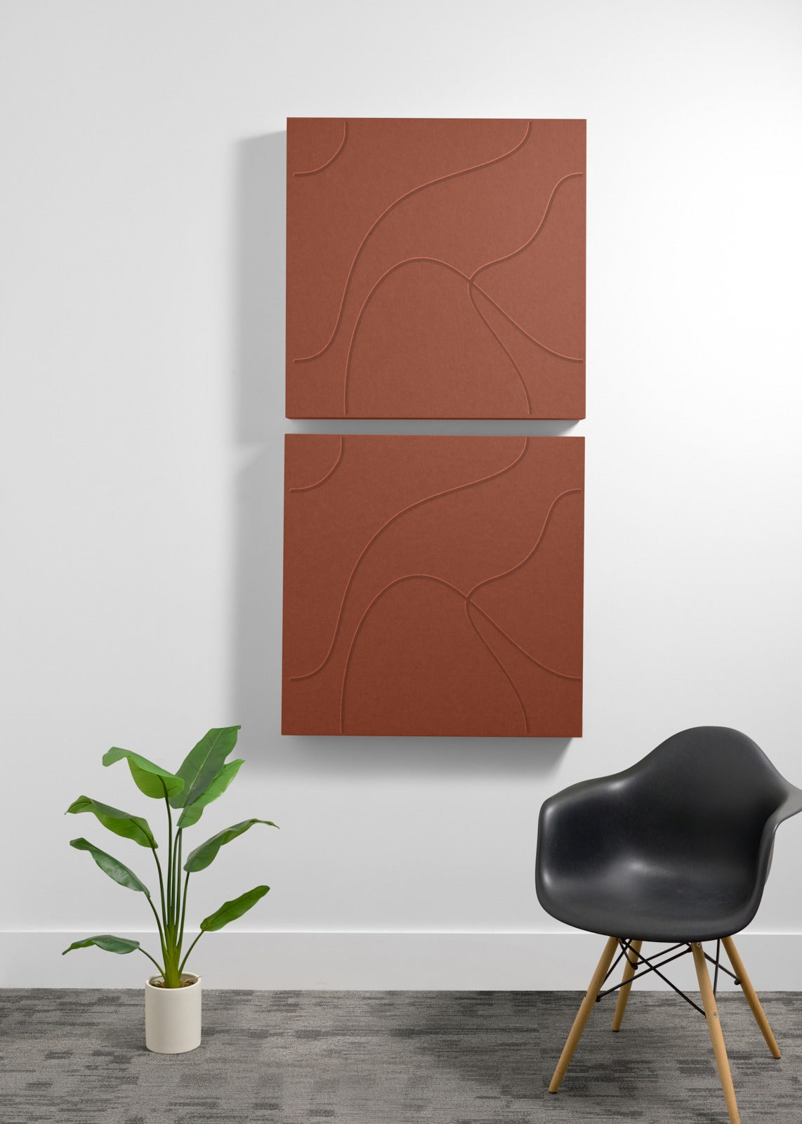 Two stacked terracotta-colored square panels with engraved curved line patterns mounted on white wall above black chair.