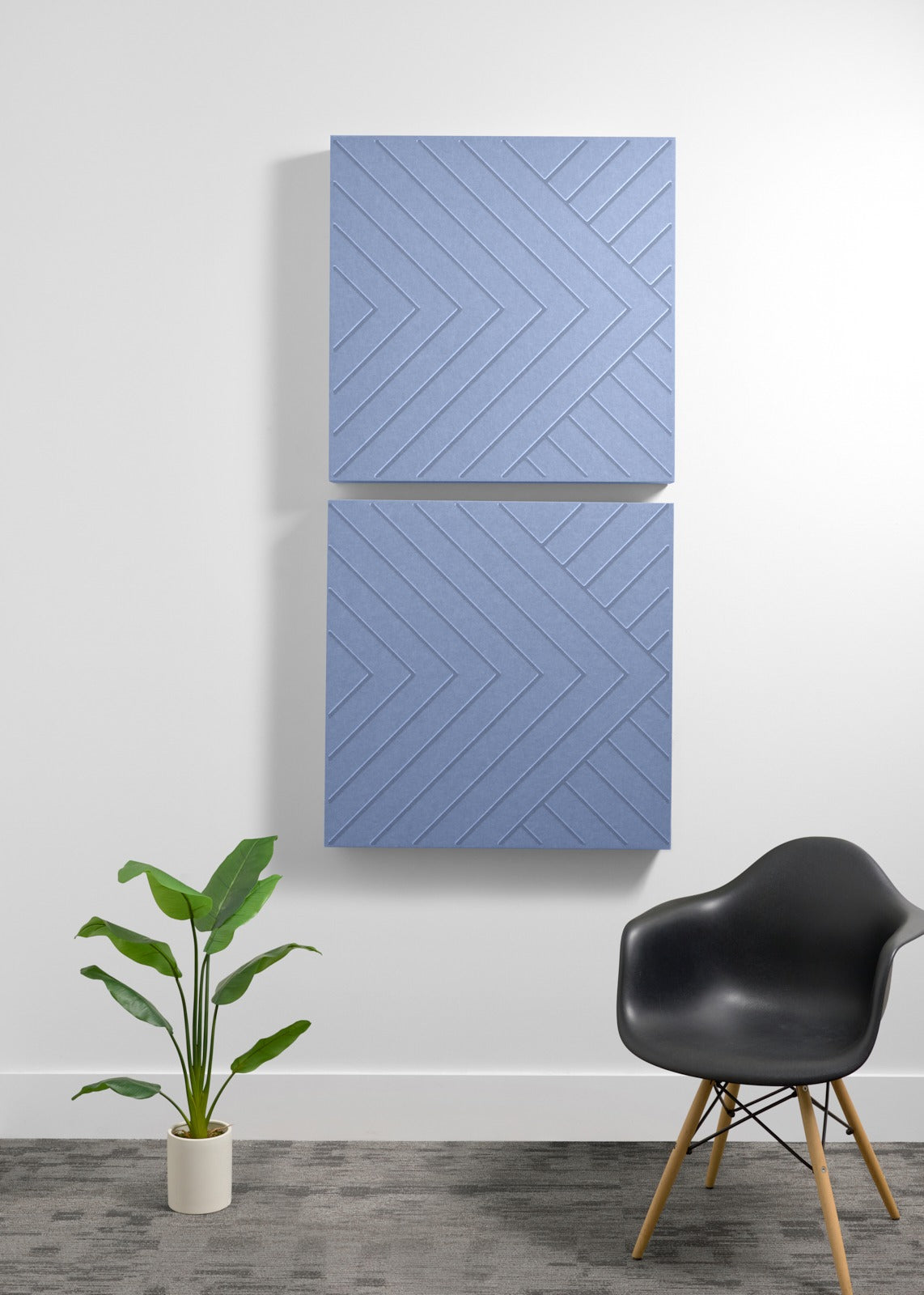 Two stacked blue acoustic panels with engraved chevron pattern mounted on wall above potted plant and black chair.