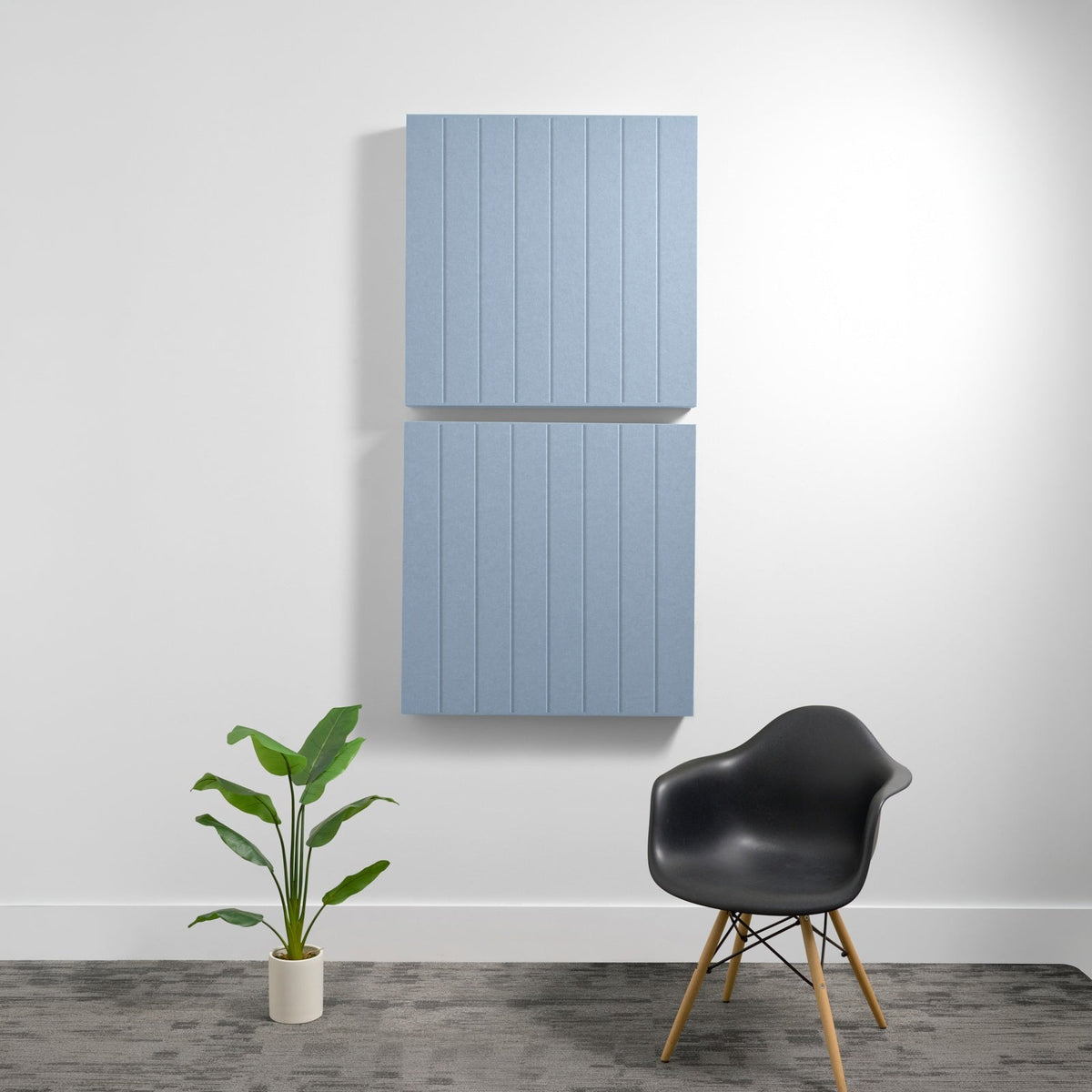 Two stacked light blue acoustic wall panels with parallel vertical engraving, mounted on white wall above black chair and pot