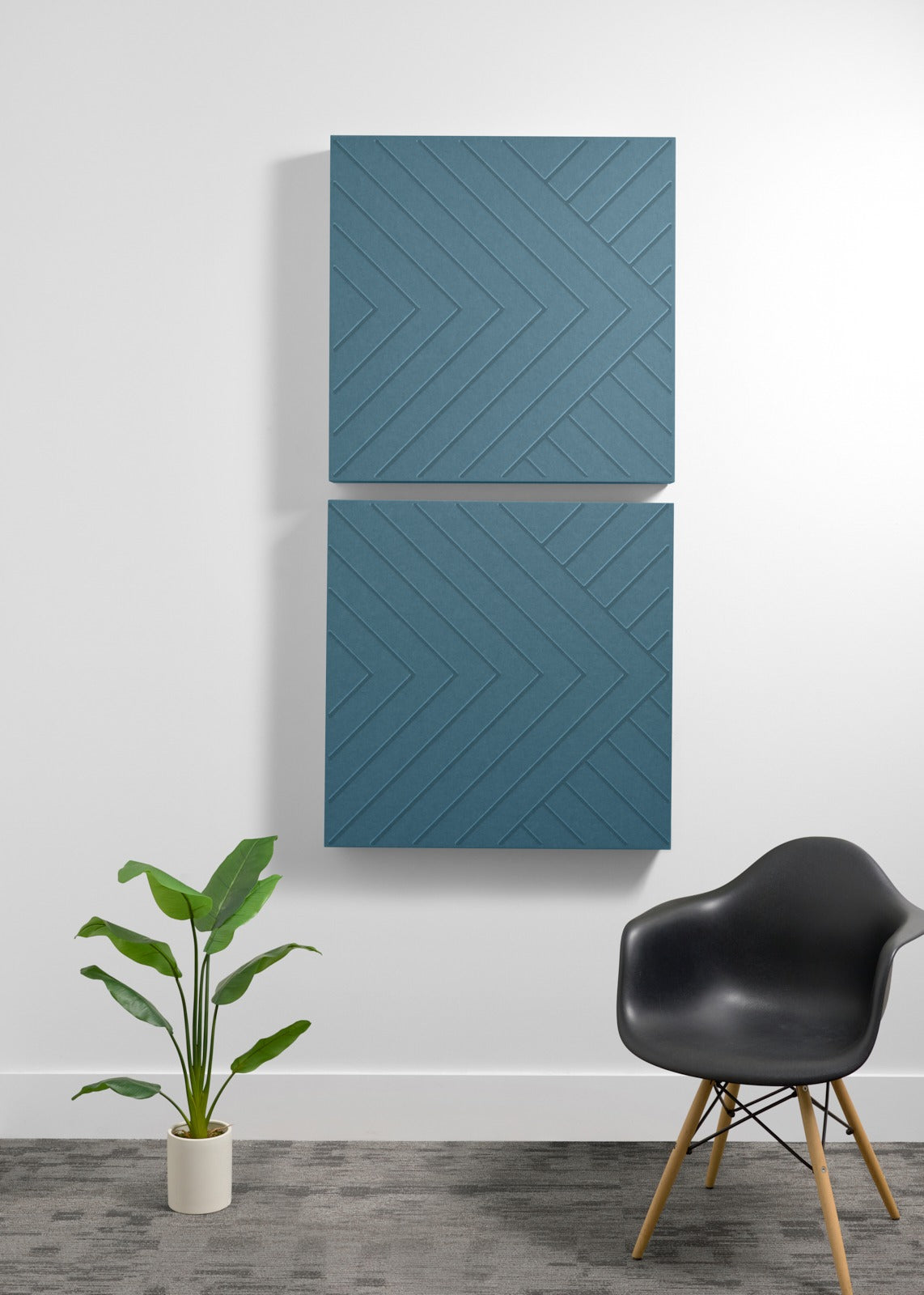 Two blue-gray square acoustic panels with geometric chevron engraving mounted on wall above chair and plant.