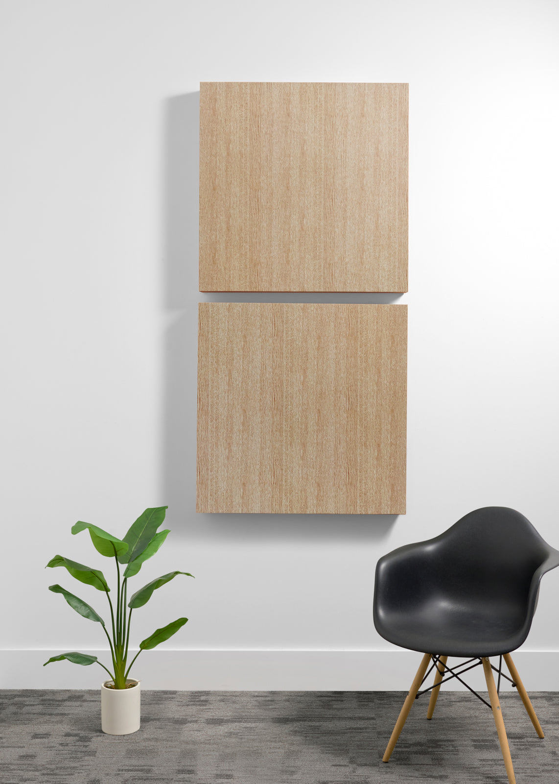 Two square light wood wall art panels hanging vertically on a white wall in a modern room setting, with a black chair and a potted green plant below.