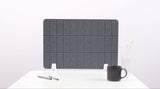 Gray padded desk divider with grid pattern and white mounting brackets on white desk with coffee mug and pens.