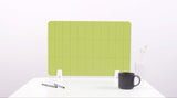 Lime green grid desk divider with white mounting brackets, positioned on white desk with pens, black mug, and vase.