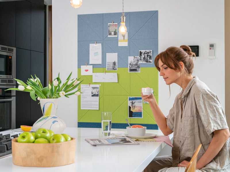 a felt right wall art design in a kitchen with blue and green felt tiles creating a pinable pattern on a white wall with a woman drinker her coffee reading a paper.
