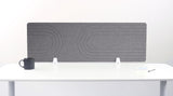 Gray desk divider with geometric line patterns, standing on white desk supports between office supplies.
