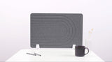 Gray desk divider with curved linear pattern design, positioned upright on white desk with support feet.