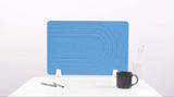 Blue desk divider with curved line pattern and white stand supports, displayed with pens, mug, and vase.