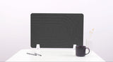 Dark gray desk divider with concentric curved lines, standing upright on white supports on a white desk with a mug and pen ho