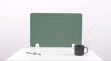 Green desk divider with curved line pattern, white stand, positioned on white desk with coffee mug and pen holder.