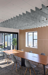 A modern conference room with a long wooden table, gray chairs, a slatted wood wall, and a large concrete staircase structure suspended from the ceiling.