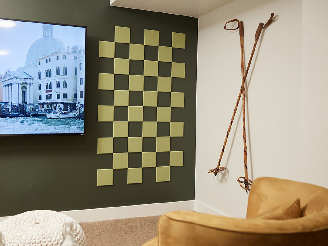 A Felt Right wall art design in a shade of green square felt tiles in a checkered pattern on a darker green wall next to a tv and ski poles on the other wall