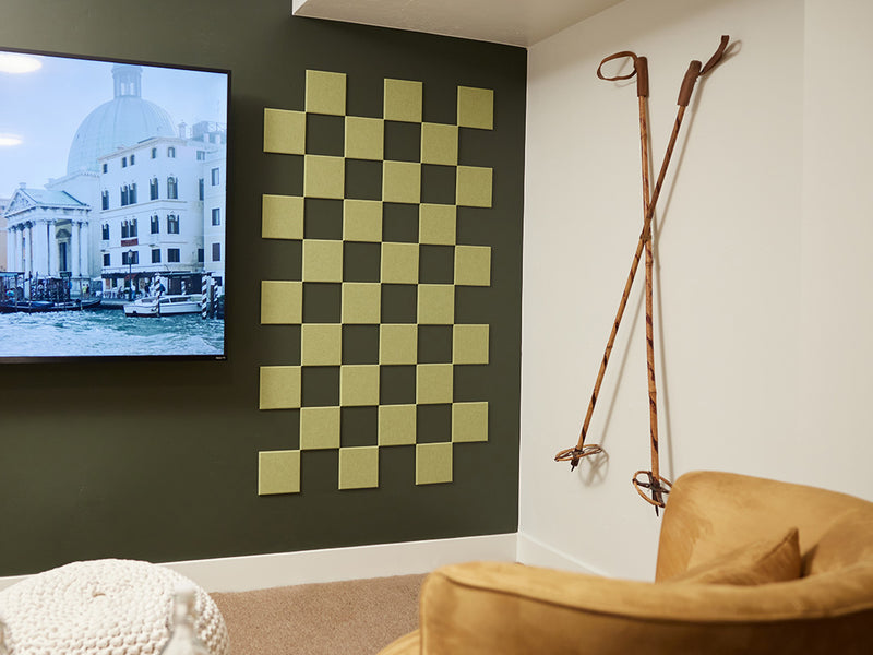 A Felt Right wall art design in a shade of green square felt tiles in a checkered pattern on a darker green wall next to a tv and ski poles on the other wall