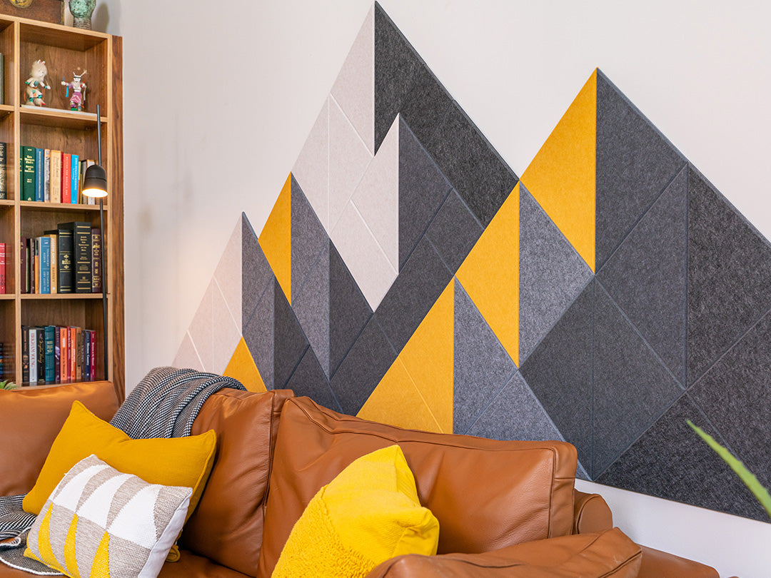 A Felt Right wall design wall decor featuring a geometric mountain-like pattern in shades of gray, black, and yellow, mounted on a wall above a brown sofa with yellow and gray throw pillows.
