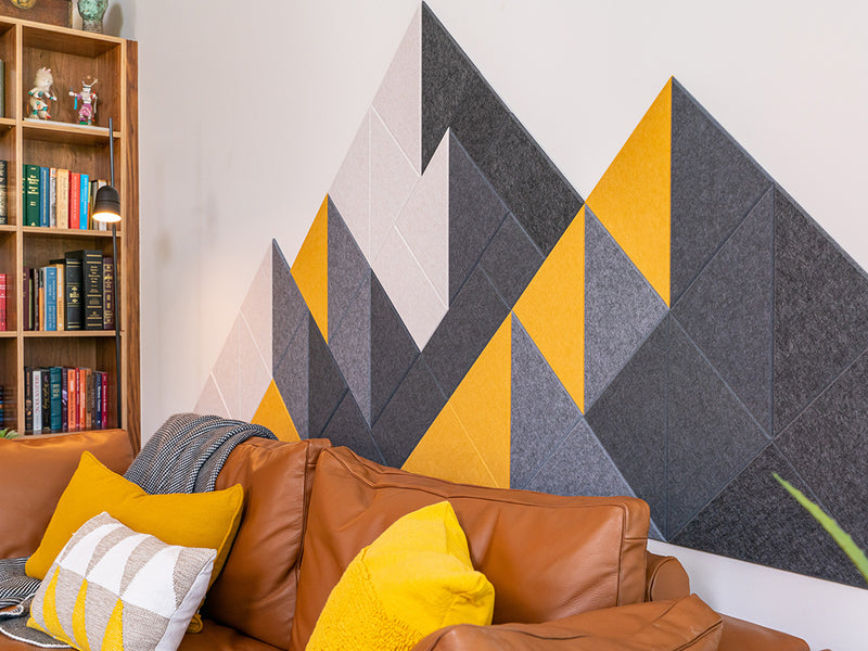 A Felt Right wall design wall decor featuring a geometric mountain-like pattern in shades of gray, black, and yellow, mounted on a wall above a brown sofa with yellow and gray throw pillows.