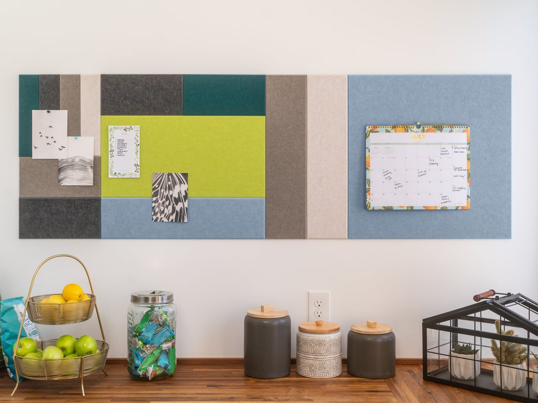 Felt Right wall art design using blue, grey, green, and cream shades of felt tiles that are pinable with pictures and a calendar above a table with fruit and jars on it.