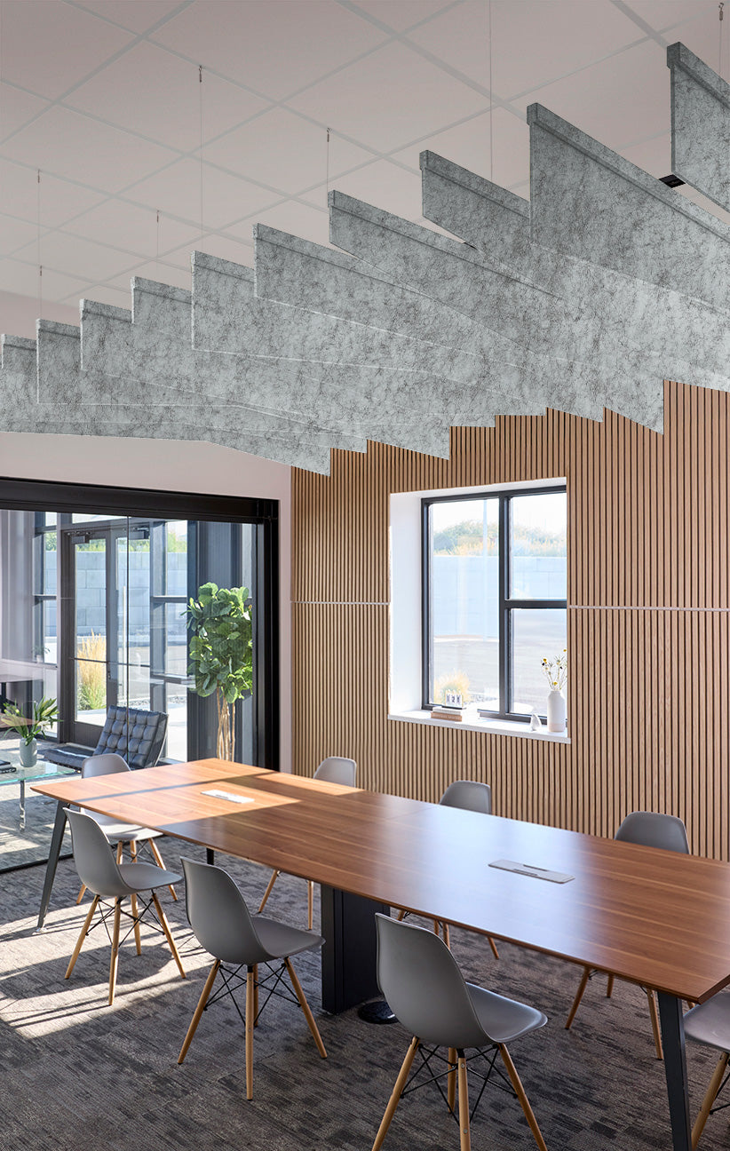 Modern conference room with a long wooden table, gray chairs, a slatted wood accent wall, large windows, and a distinctive concrete staircase sound barrier hanging from the ceiling.