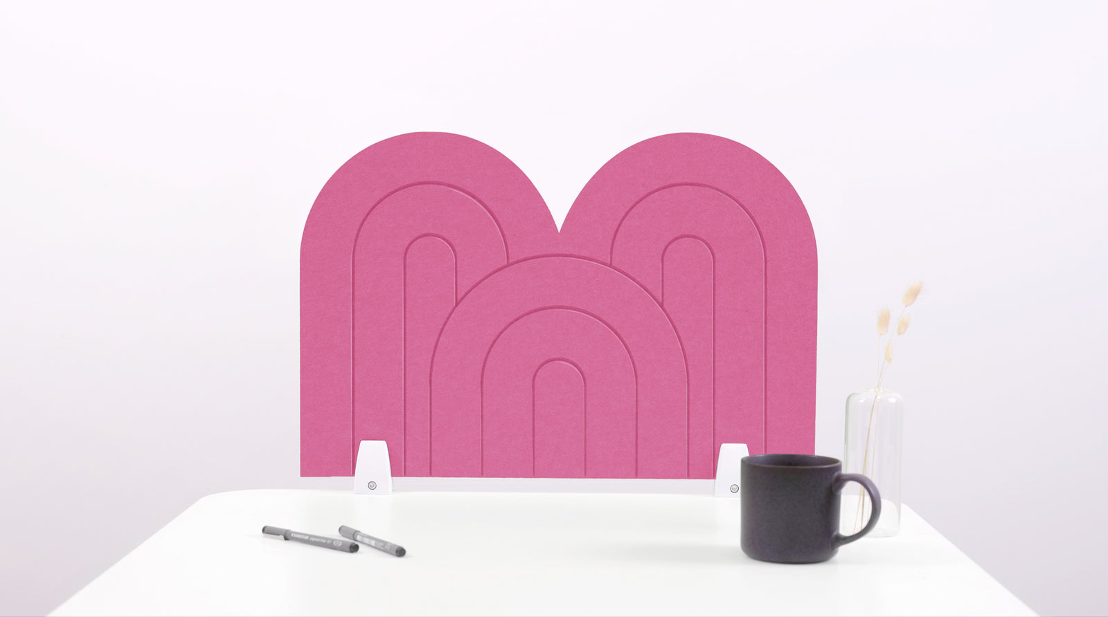 Pink arch-shaped desk divider with nested geometric patterns, displayed on white desk with black mug and pens.