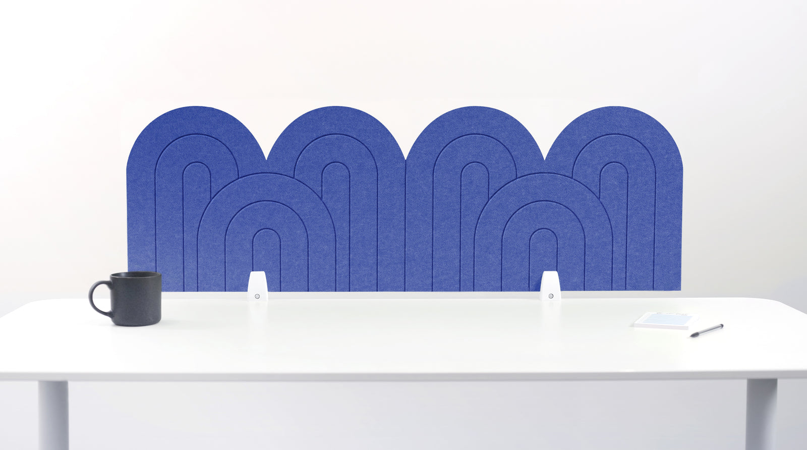 A dark blue felt desk divider with a wavy, layered arch pattern, shown standing behind a white desk with a black mug on the left and a notepad on the right.