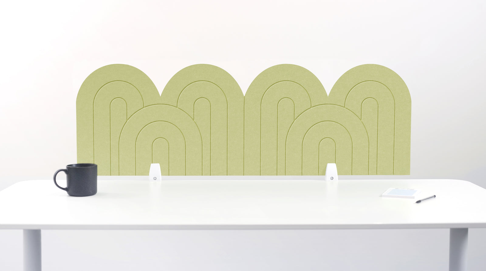 An green felt desk divider with a wavy, layered arch pattern, shown standing behind a white desk with a black mug on the left and a notepad on the right.