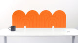 An orange felt desk divider with a wavy, layered arch pattern, shown standing behind a white desk with a black mug on the left and a notepad on the right.