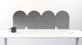 A gray felt desk divider with a wavy, layered arch pattern, shown standing behind a white desk with a black mug on the left and a notepad on the right.