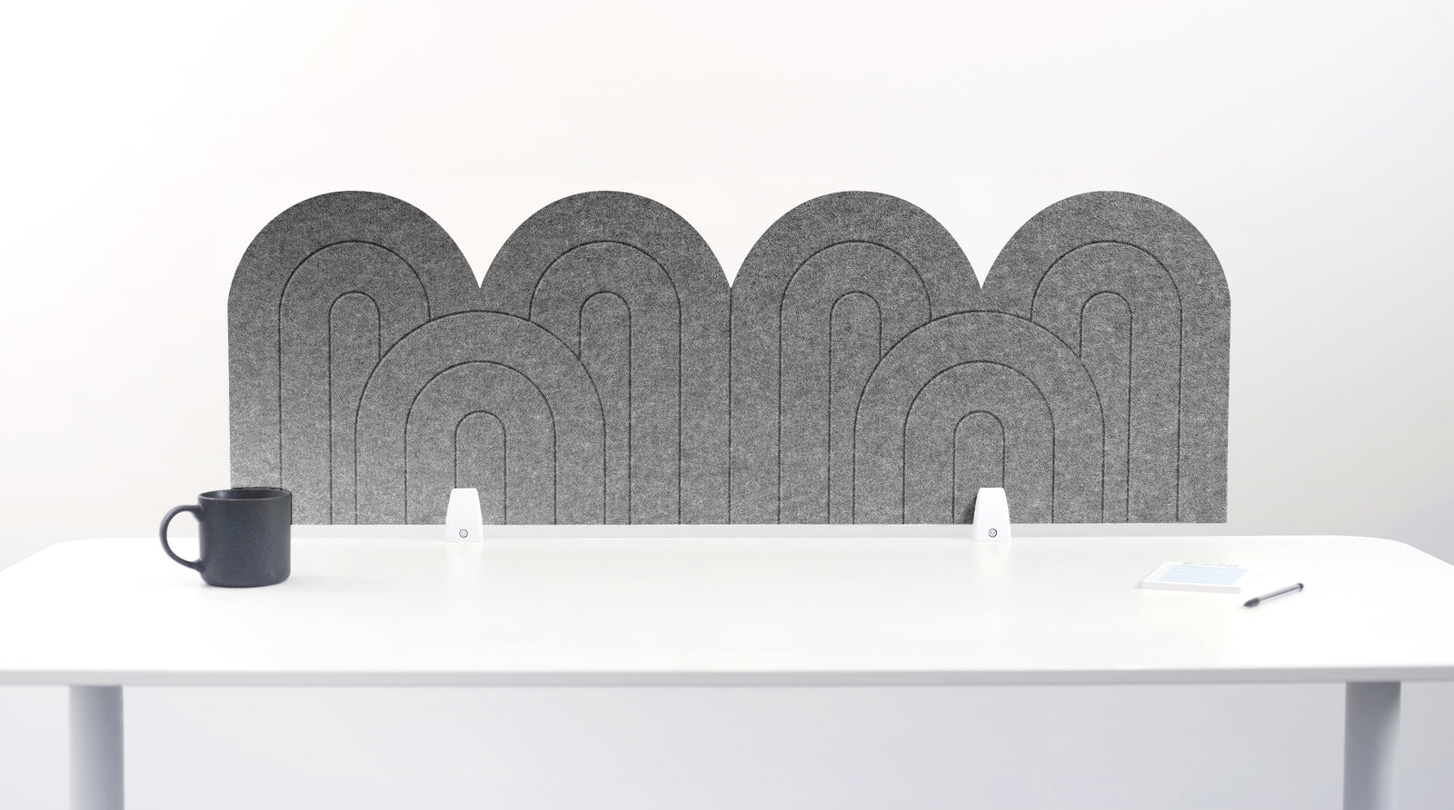 A gray felt desk divider with a wavy, layered arch pattern, shown standing behind a white desk with a black mug on the left and a notepad on the right.