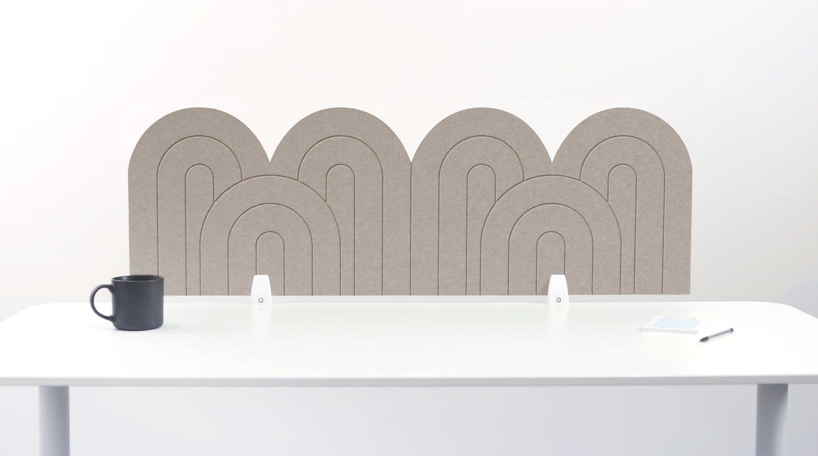 A long ash colored felt desk divider with a wavy, layered arch pattern, shown standing behind a white desk with a black mug on the left and a notepad on the right.