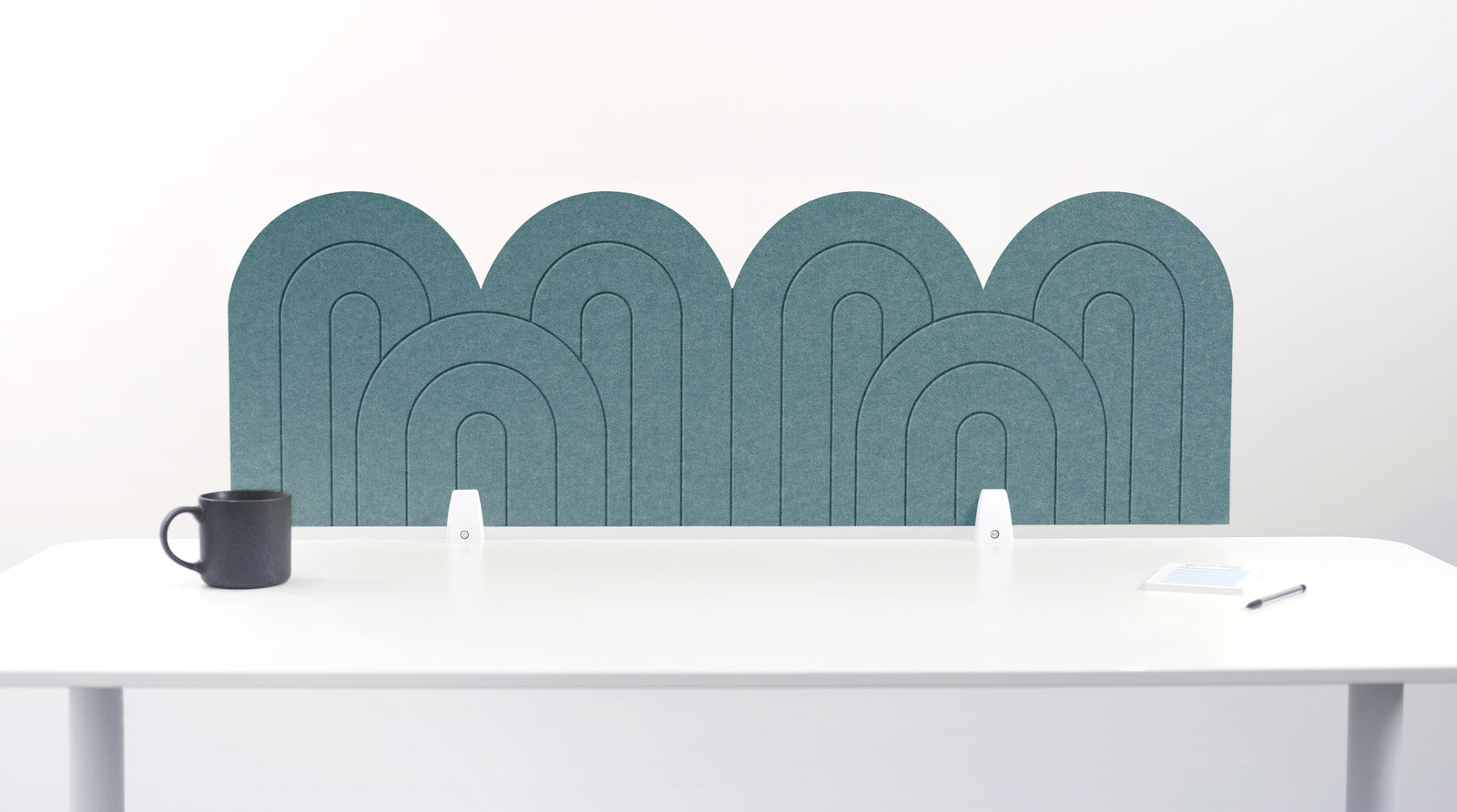 A teal blue felt desk divider with a wavy, layered arch pattern, shown standing behind a white desk with a black mug on the left and a notepad on the right.