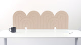 A light tan felt desk divider with a wavy, layered arch pattern, shown standing behind a white desk with a black mug on the left and a notepad on the right.
